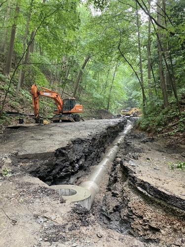Two excavators operate on a steep road through green forest. The asphalt has already been removed. On the right edge is a trench with a new storm sewer pipe.