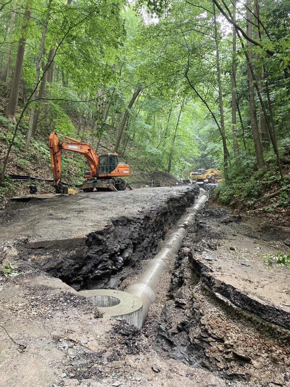 Two excavators operate on a steep road through green forest. The asphalt has already been removed. On the right edge is a trench with a new storm sewer pipe.