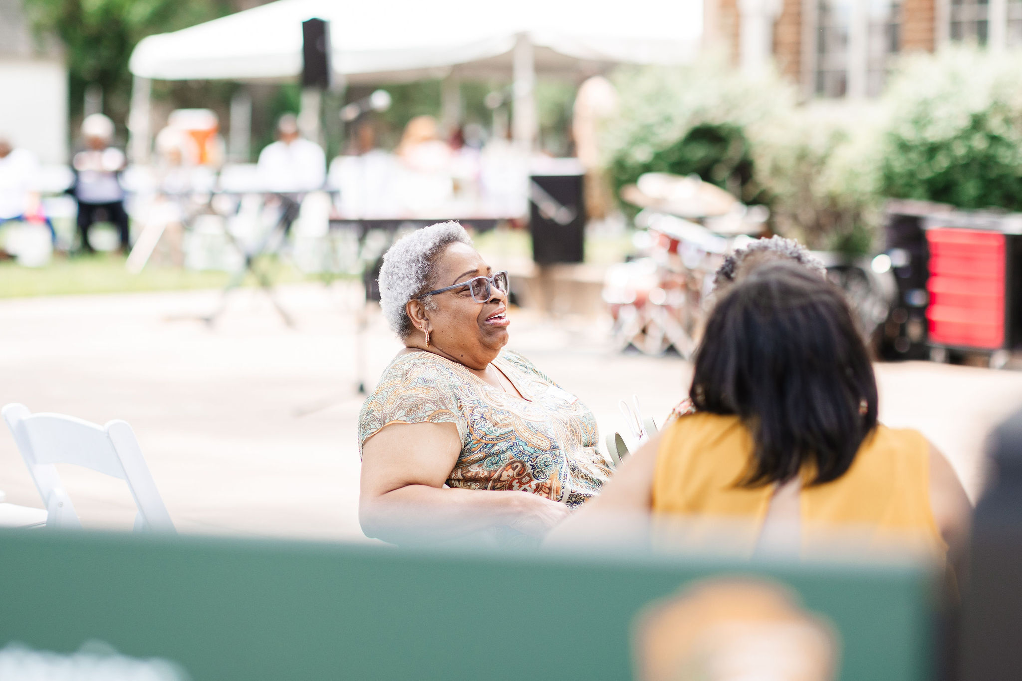An older African American woman in a paisley patterned shirt leans back and smiles while watching the community performances for the Homecoming Celebration which were held infront of Monroe Elementary.