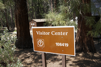 A brown metal sign stands in front of two trees, behind the trees there are wooden buildings. 