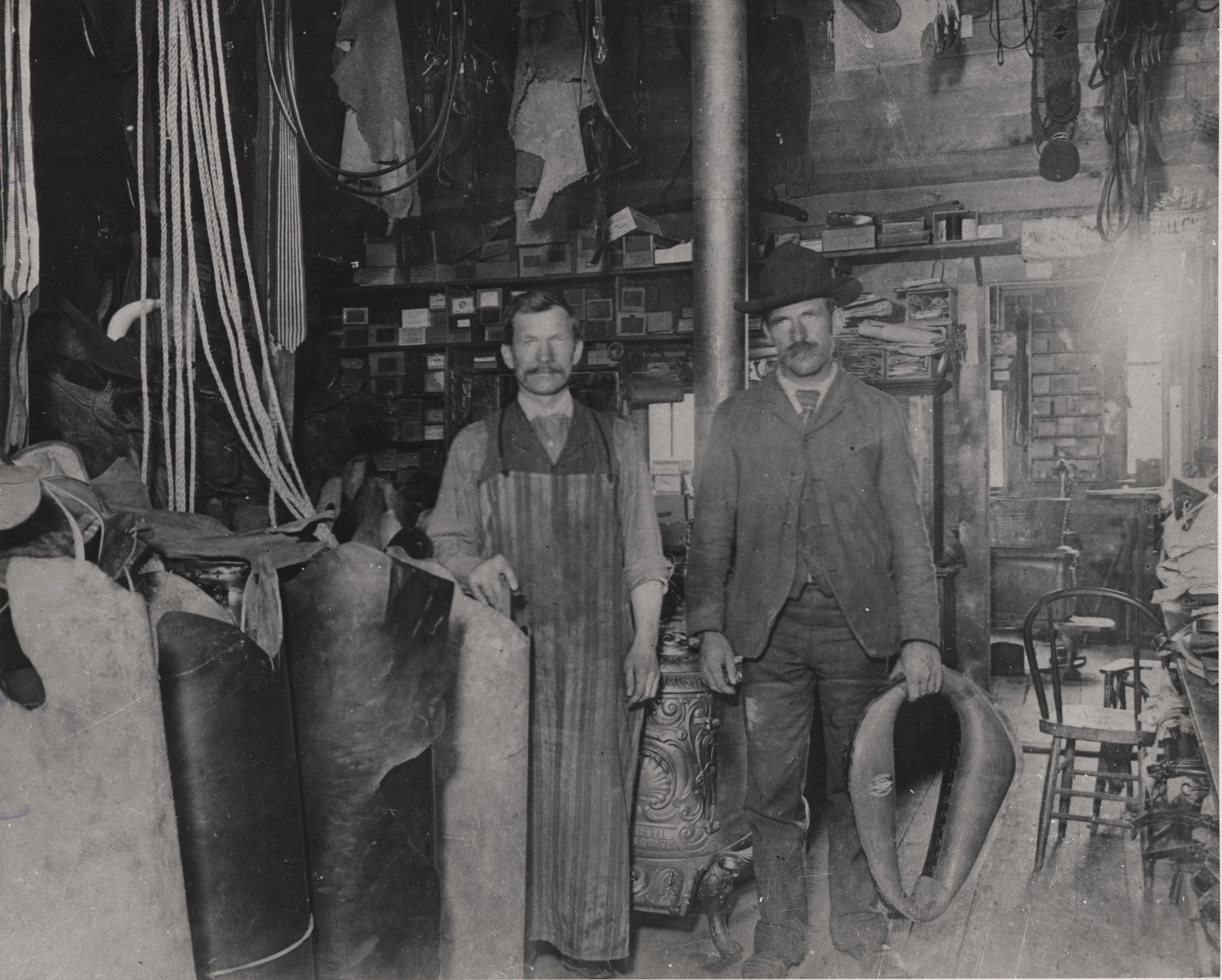 Black and white photograph of two men standing inside a store, surrounded by rope, leather, and many shelves and boxes.