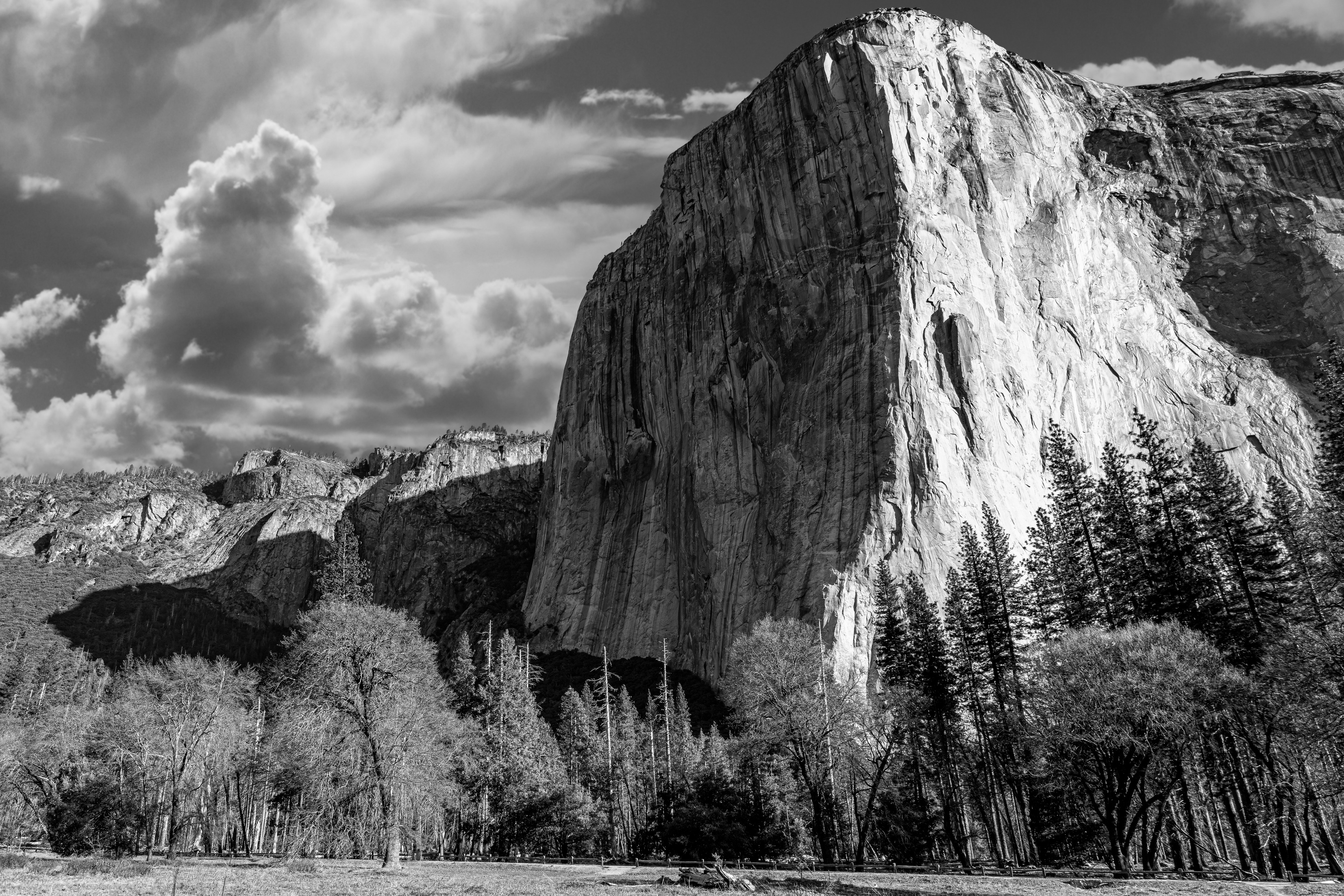 Black and white high contrast photo of a large granite rock formation along a mountain range. Trees in foreground and clouds in background.