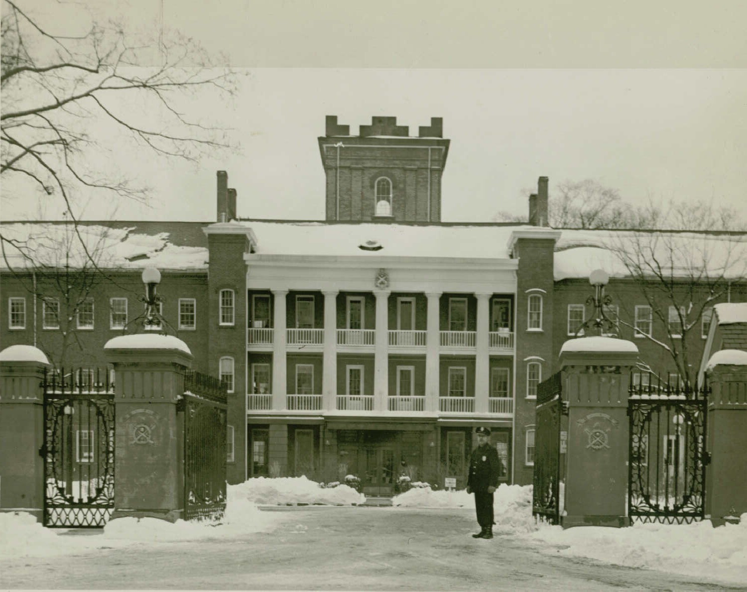 Black and white photo of part of the front of a large brick building with a second and third story balcony and a tower in the back. An insignia hangs above the middle of the third story balcony. A metal gate with stone pillars is open in the foreground. A man stands in the open gate on the right. Light snow covers the ground, roof, and pillars and is piled to the side of the driveway and sidewalk.