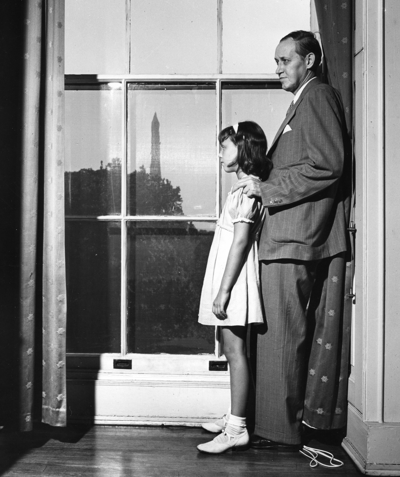 A man in a suit standing by a window with a young girl in a dress. The Washington monument is in the background.