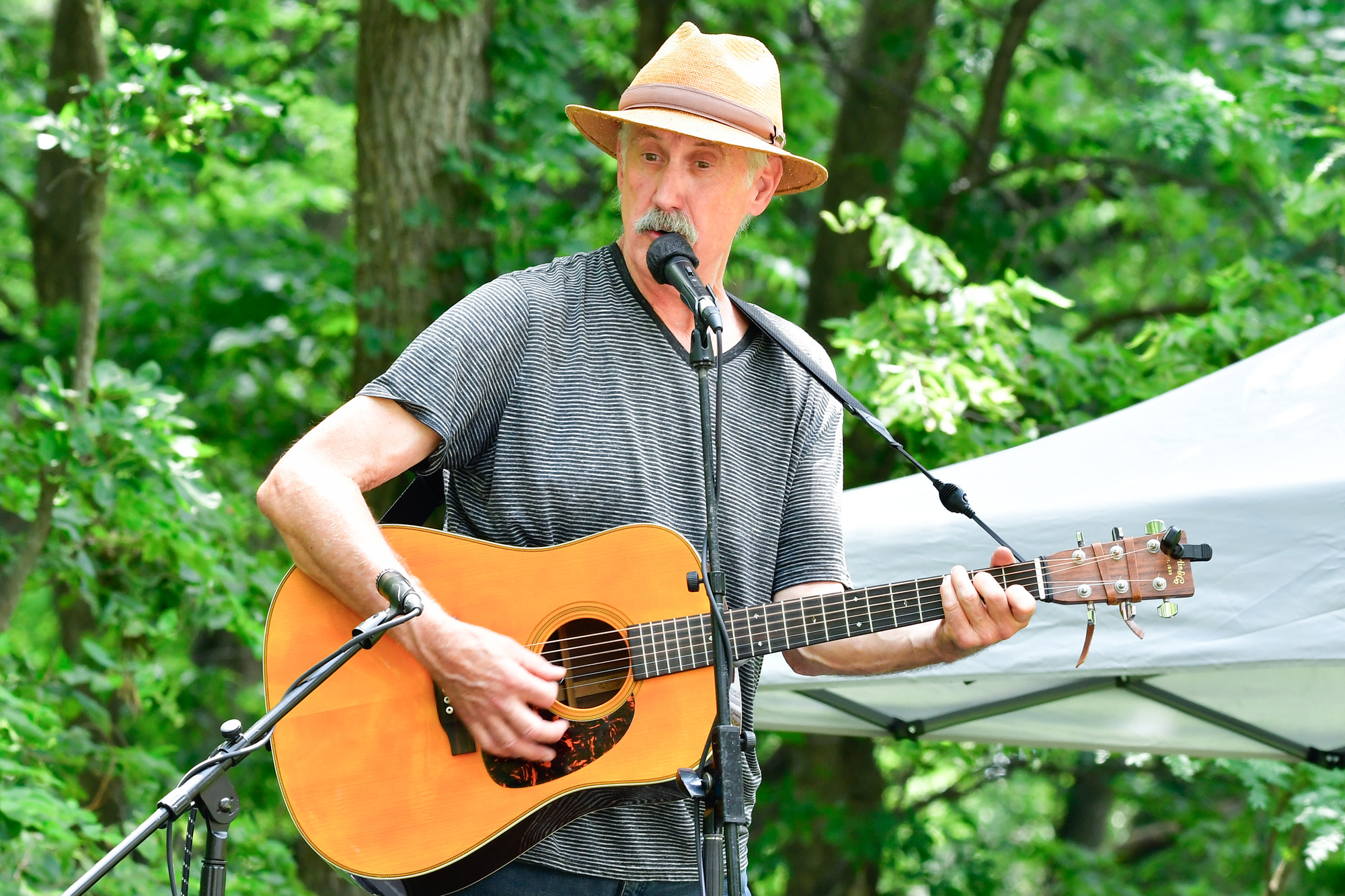 Man in a black and white striped shirt and straw hat plays guitar and sings into a microphone.