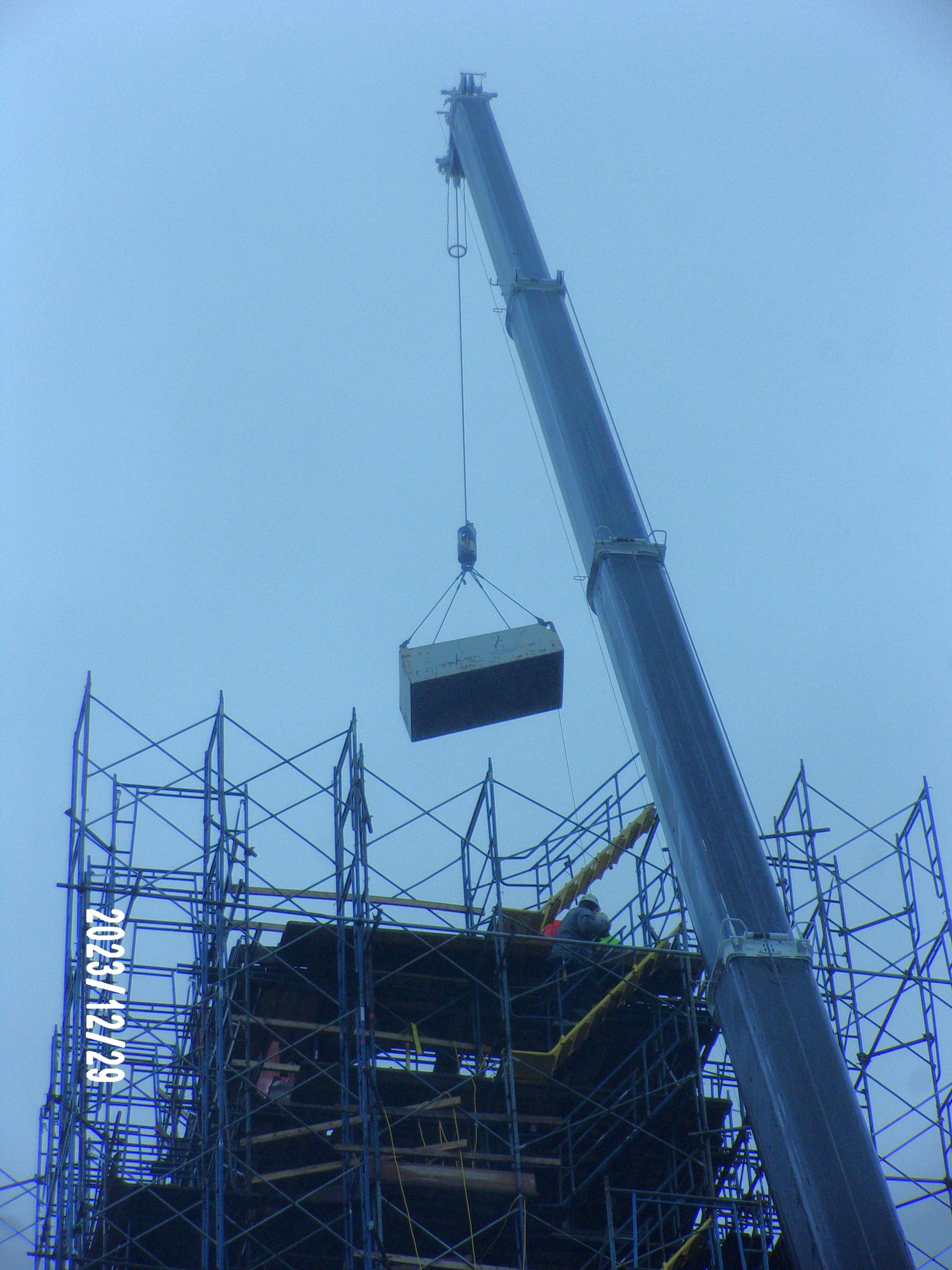 Against a foggy sky a crane hoists a rectangular container approximately 6 feet by 3 feet and 2 feet deep from a monument shrouded in extensive scaffolding