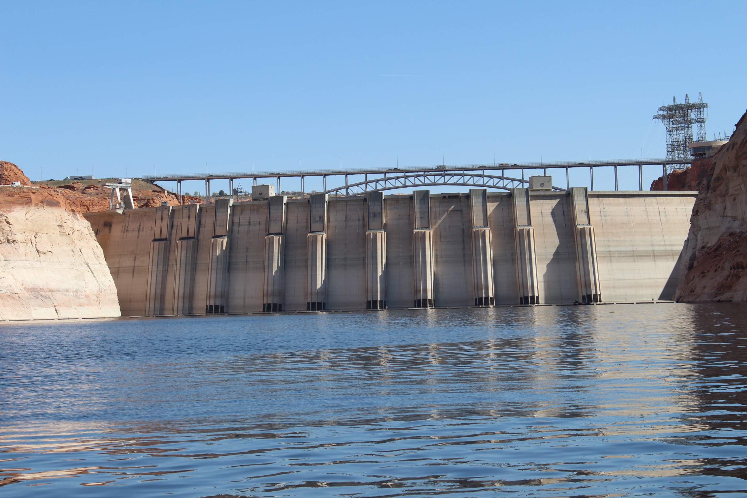 Concrete structure holding back water in steep rock canyon. A metal suspension bridge with vehicles rises above the top of the structure. 