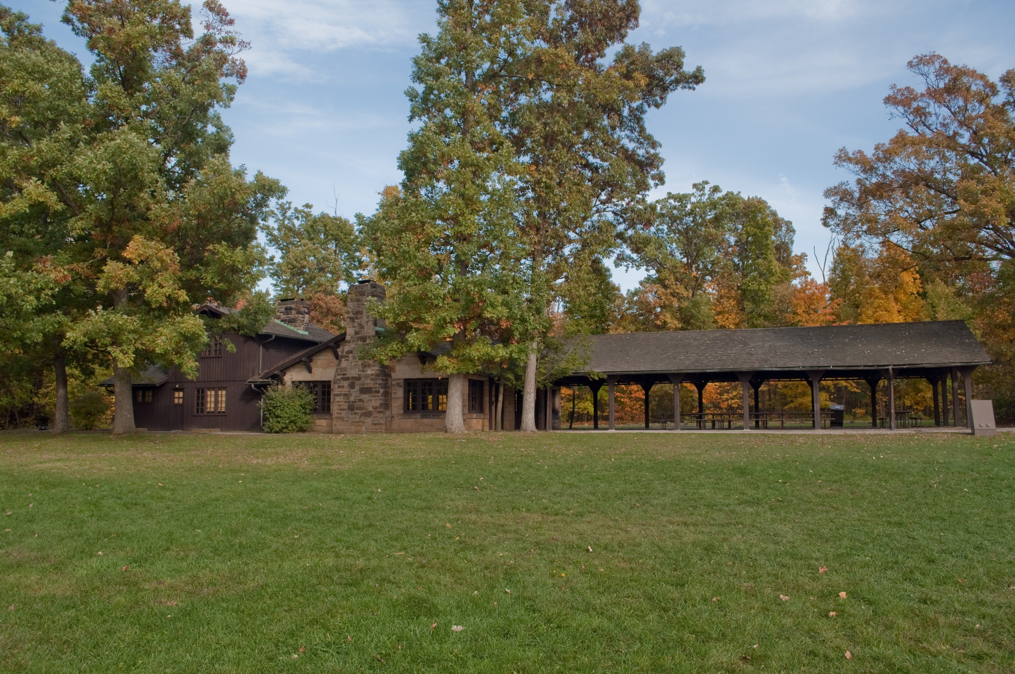 Side view of a two story brown wood and gray stone building; the right side is a one-story covered porch with brown columns; tall trees grow along its sides and in the background and mowed grass in the foreground.