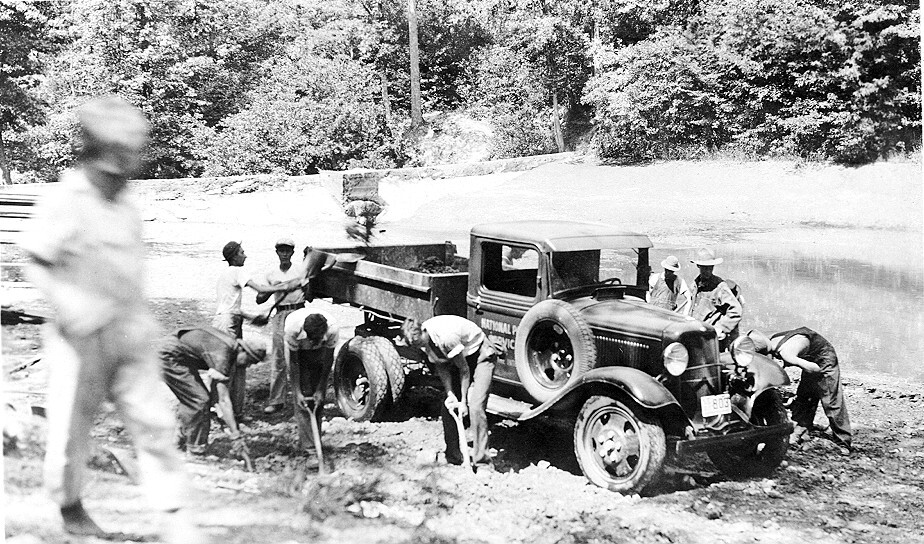 A group of young men are working on a piece of road. Some are digging into the ground with shovels while others are dumping dirt into the back of an old  truck.