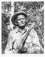 Historic black and white photo of a man with a sour look holding an axe outside.