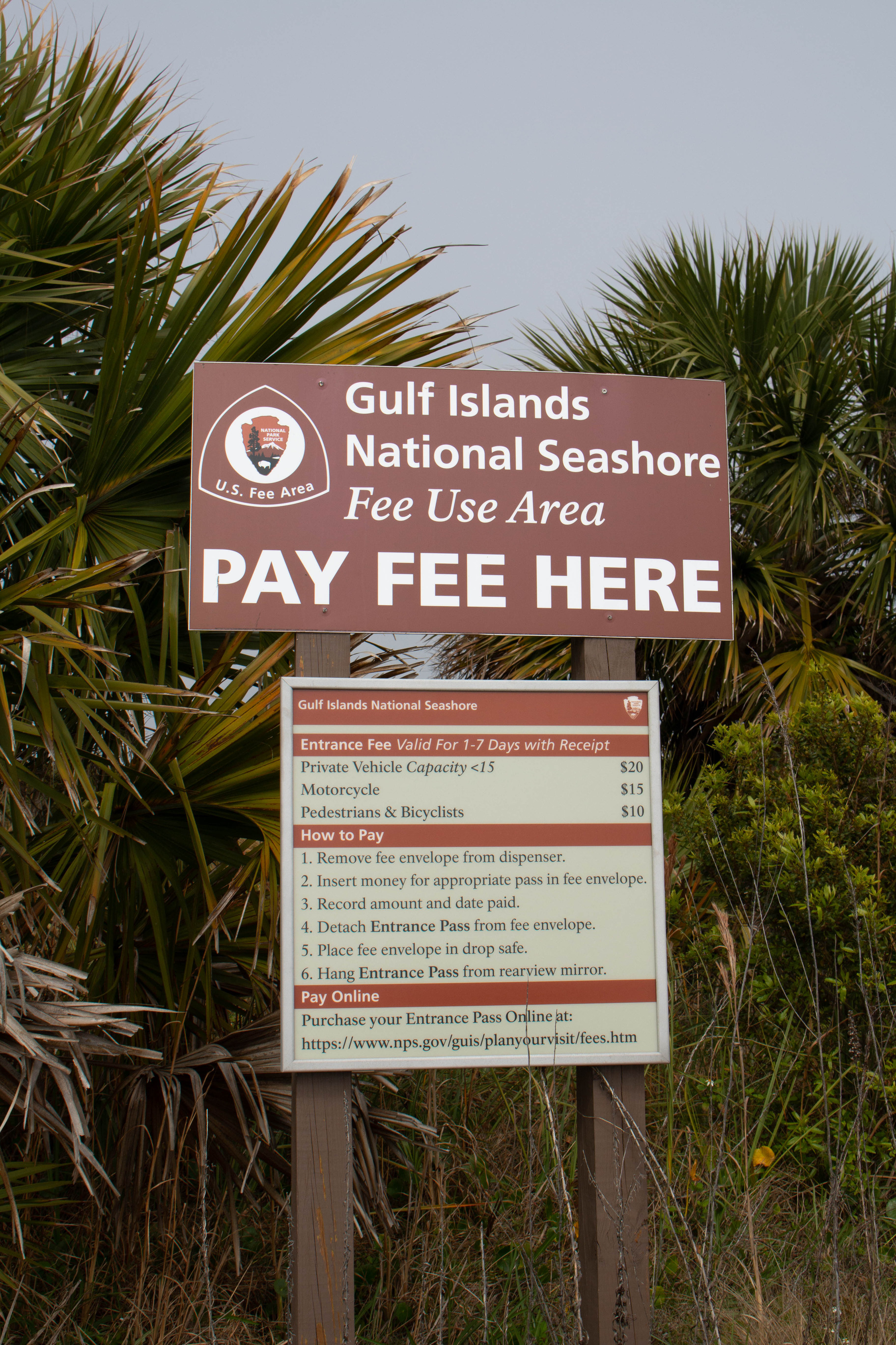 Brown metal sign with white, "Fee Use Area, Pay Fee Here" text. Palm trees in the background.