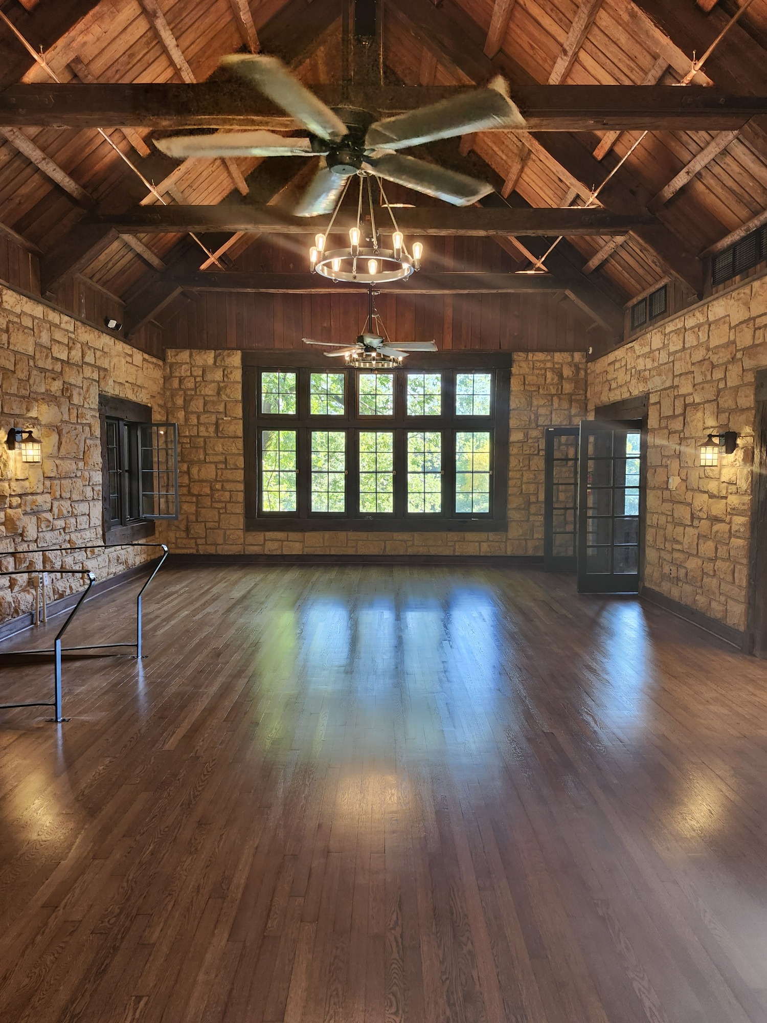 An open dining room with table settings cleared out. The floor is newly finished hardwood, the walls are coursed-cut stone blocks with a large bank of windows at the far end, and the ceiling is peaked and made from boards running from bottom to top. The scene is illuminated with warm light by sconces and chandeliers.
