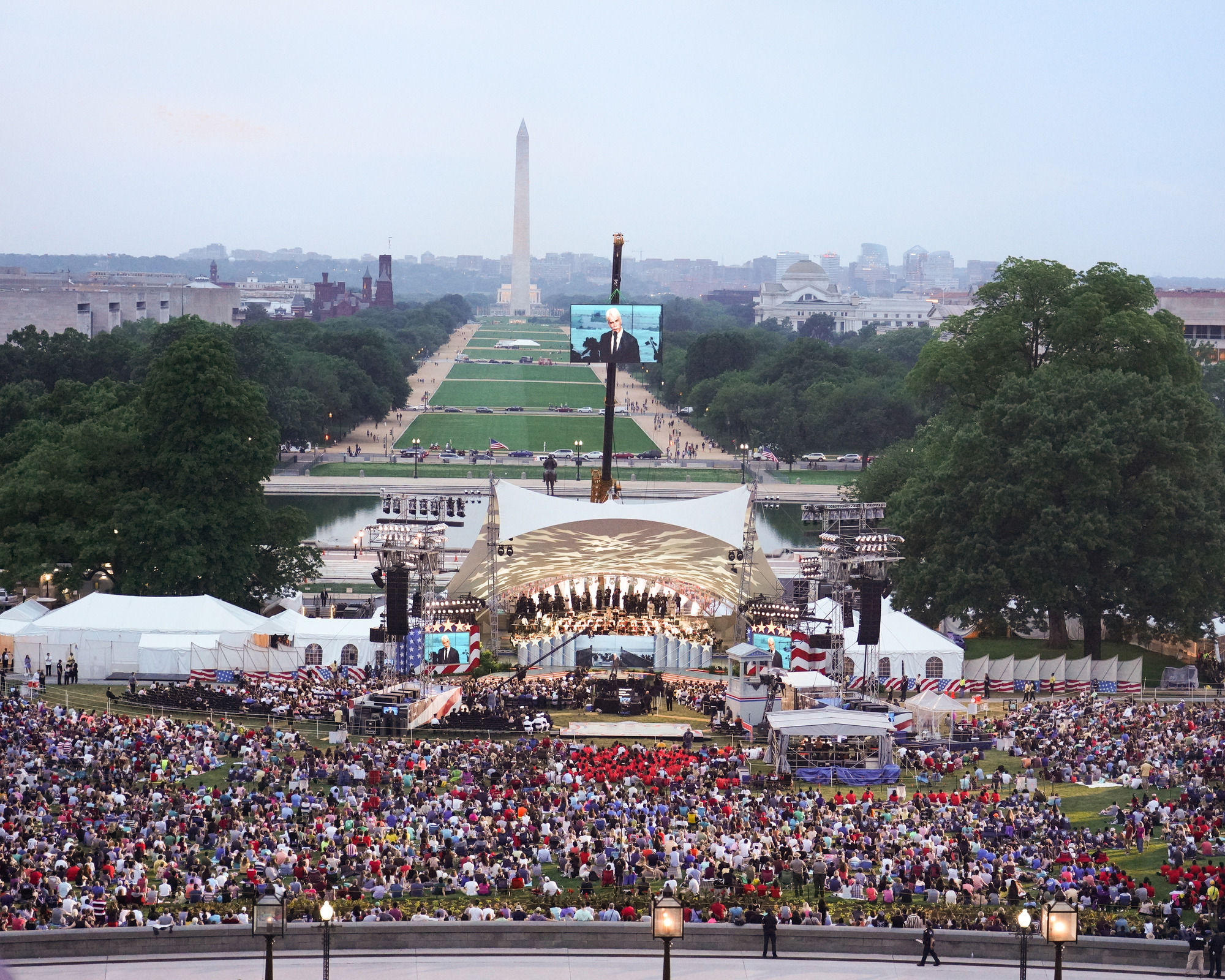 A crowd gathers in front of a bandshell with the National Mall and Washington Monument in the background.