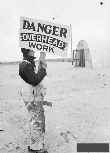 Black and white photo of a man in a sweater and overalls holding up a sign that reads "DANGER OVERHEAD WORK".