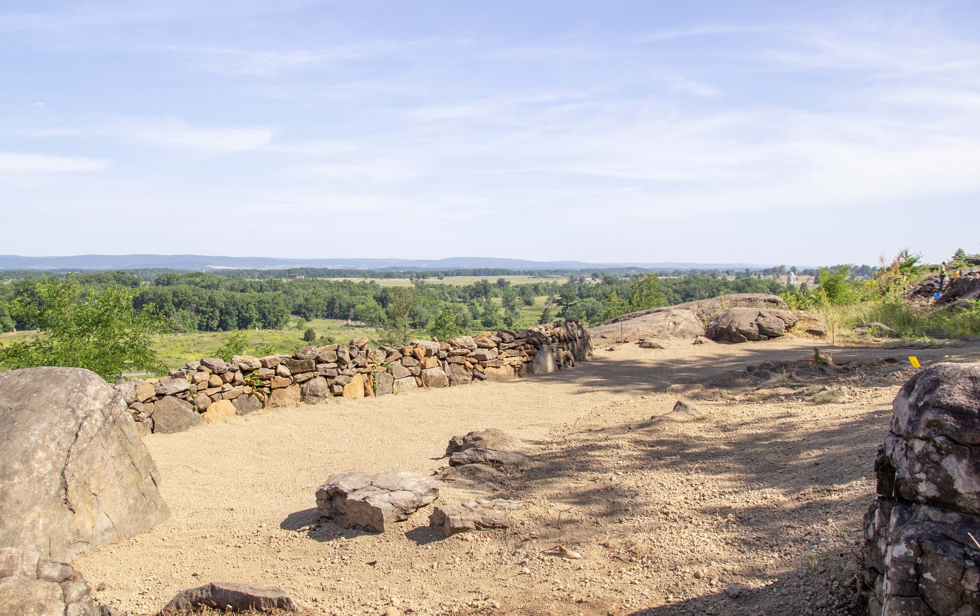 An unfinished brown stabilized aggregate gathering area sits to the right of a stone wall, and brown topsoil sits to the right of the gathering area, with large boulders and trees in the background.
