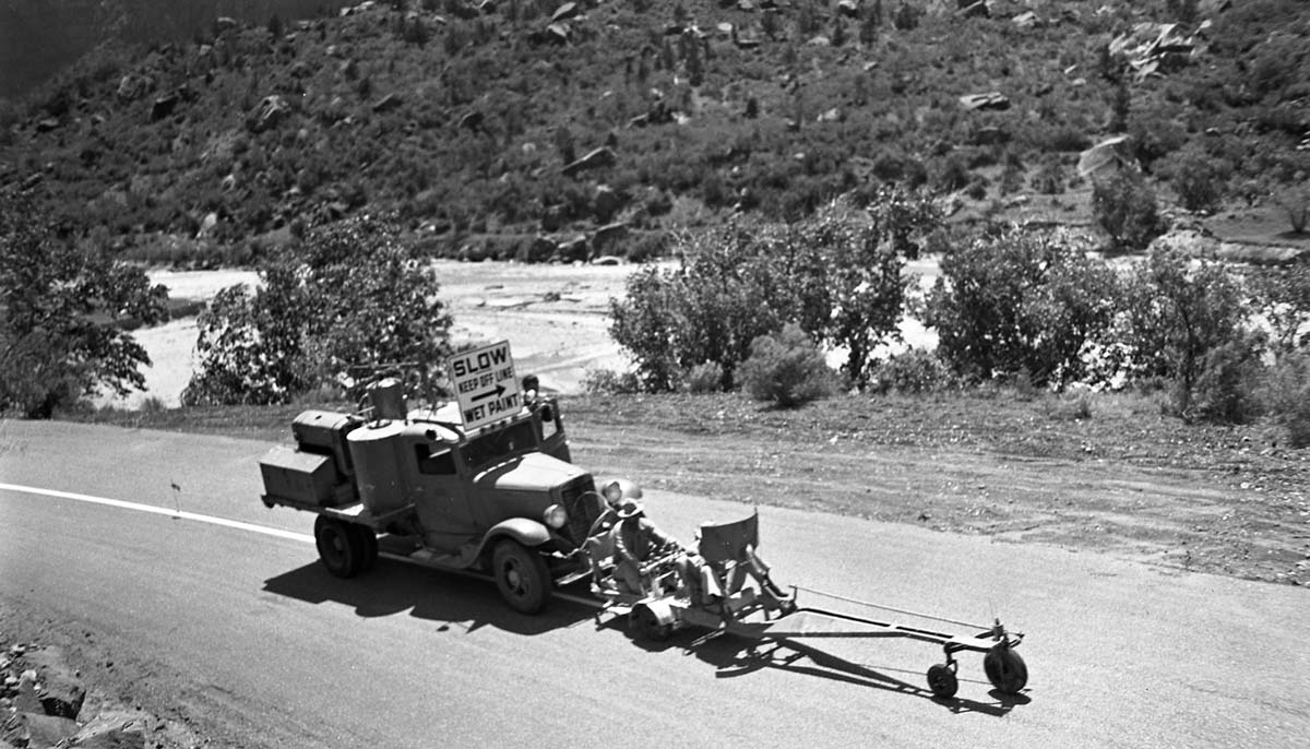 Three men operate truck and machine for painting center line stripe on road in Zion Canyon. Sign of truck says: Slow / Keep Off Line / Wet Paint.