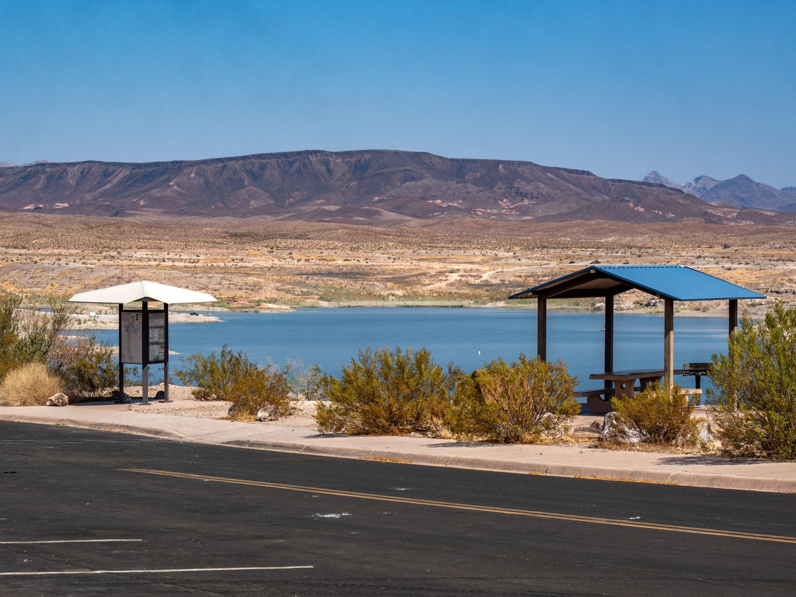 information kiosk at left, shade cover and picnic bench at right, lake and mountains behind