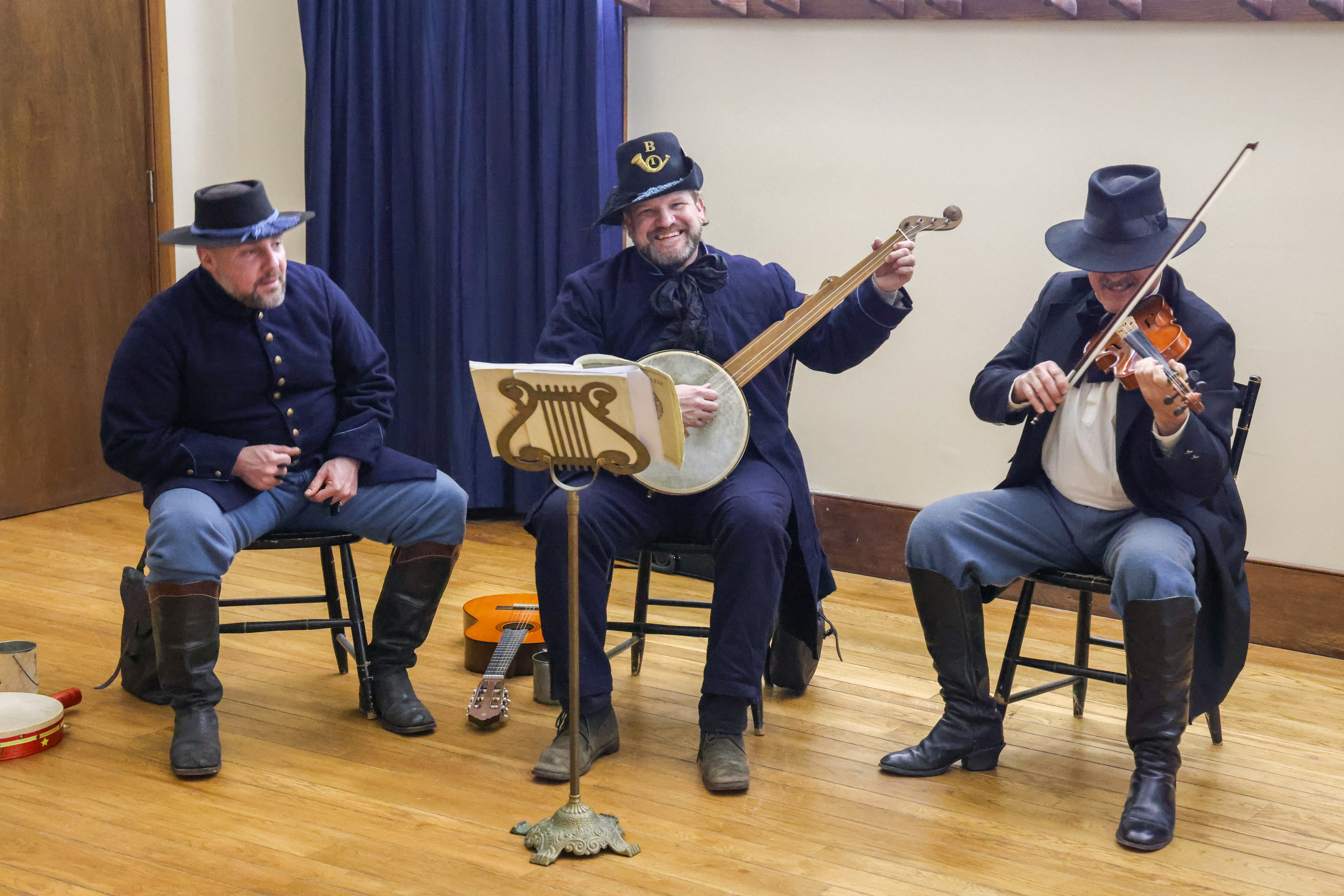 Three men wearing dark blue historic military clothing are arranged to play music. One plays a fiddle, another a banjo, and a guitar and tambourine lie ready for use next to the third man. The musician in the middle smiles and looks toward the camera.
