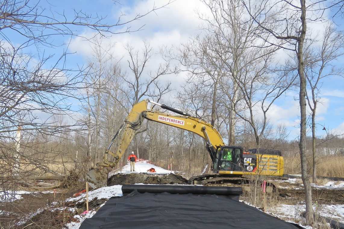 An excavator carves a deep cut that divides the snowy trail and connects the wetlands on either side. Black construction fabric unrolls in the foreground. 