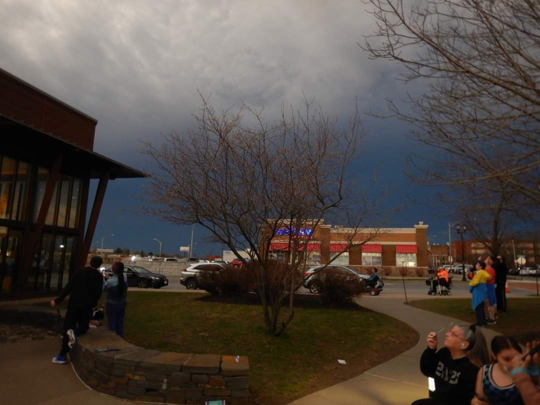 A busy scene of people in the pavillion of the park visitor center. They are looking up at the sky while a dark shadow envelopes them.