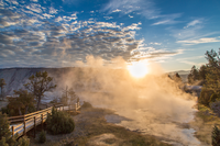 Looking at a sunrise with one person standing on a boardwalk looking at steaming hot springs .