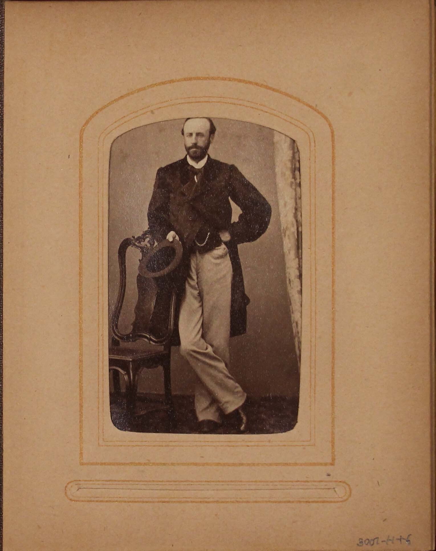 Black and white photograph of man leaning against chair holding hat.