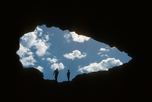 photo looking through a large arrowhead shaped hole in a dark cave ceiling at two visitors silhouetted against blue sky with clouds