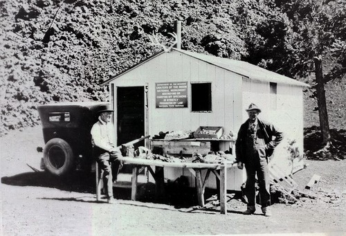 two men standing next to a table filled with rocks and artifacts in front of a small, white cabin and model T car