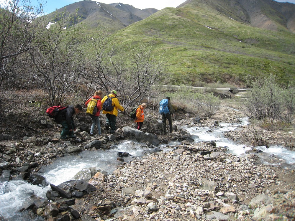 teens walk down a shalow creek, green hills in the distance