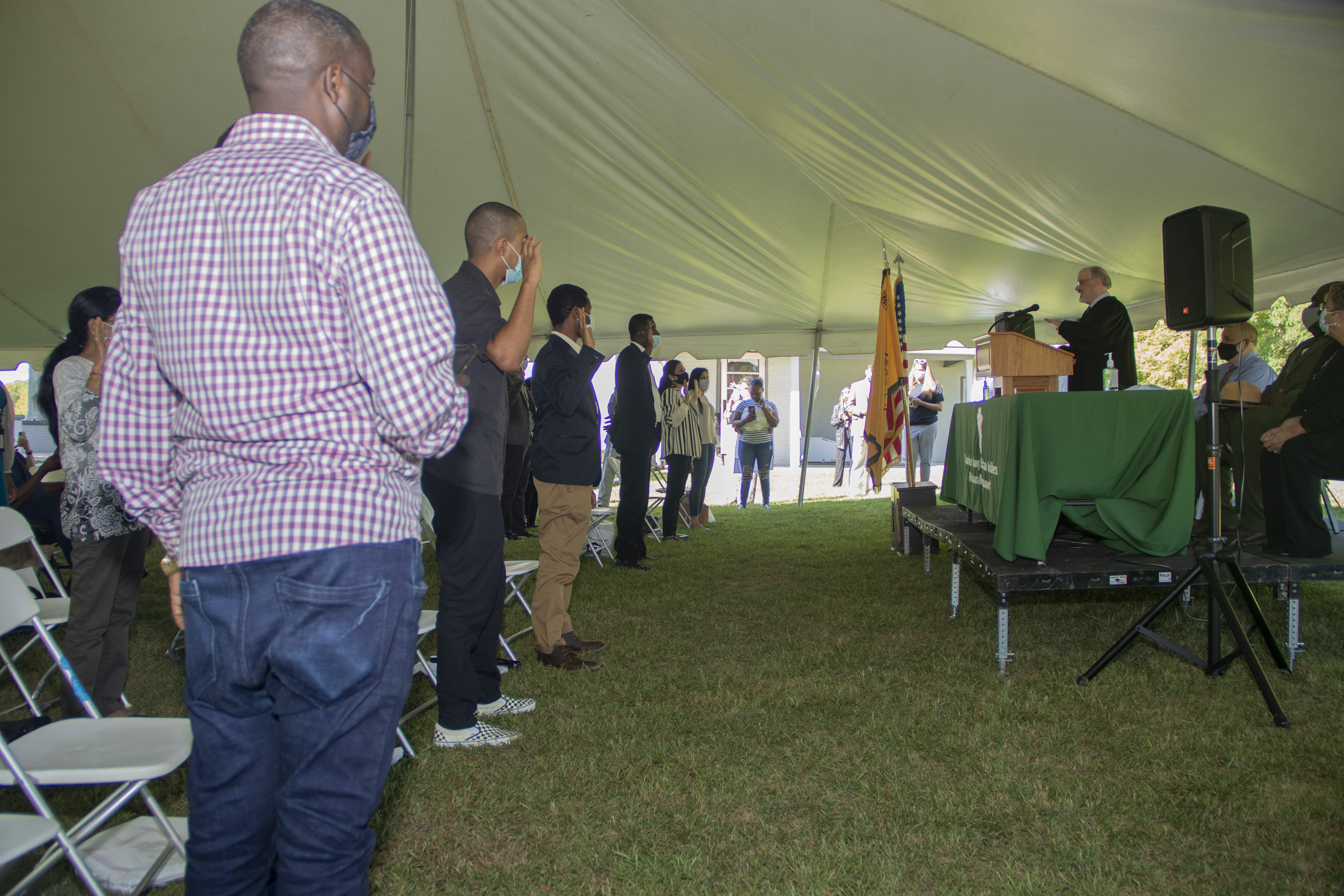 Several people standing and holding their right hands up in the air at shoulder height while two people sitting in chairs and one standing behind a podium face them from an elevated black stage behind a green-covered table.
