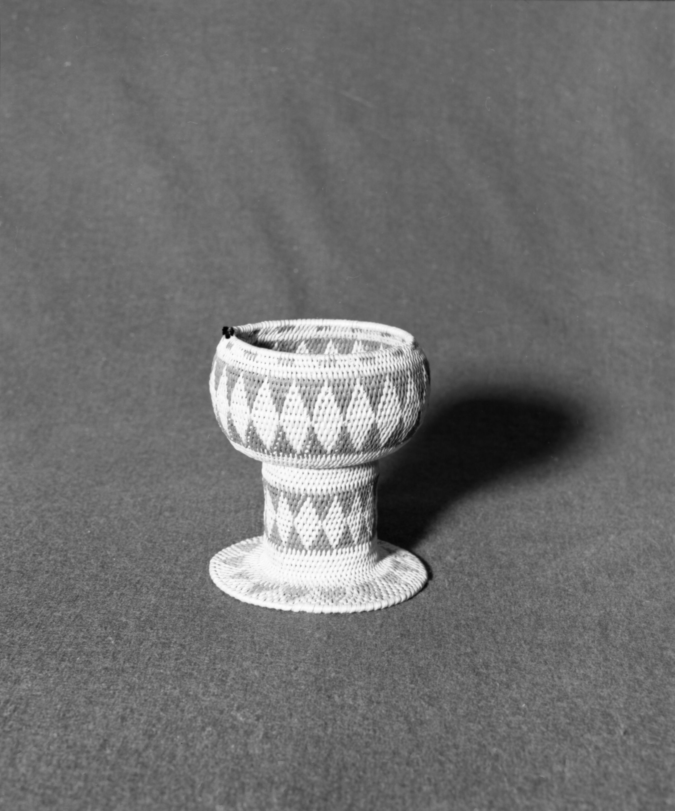 Marie Rozier Collection, Catalog No. 46. Object: Miwok pedestal basket. Dimensions: 5 3/4 x 4 1/2". Materials: fern root, bullpine. Technology: single rod, excellent example of "for sale" basket with traditional Miwok pattern, fag ends trimmed on workface, moving ends trimmed on reverse, remnants of cotton thread on rim, possibly once a handle. Collection History: From J. Frank Smith House, possibly made around 1900. Photographed by Michael Dixon, December 1982. Catalogued by Craig Bates and Barbara Beroza. Yosemite Collections, National Park Service.
