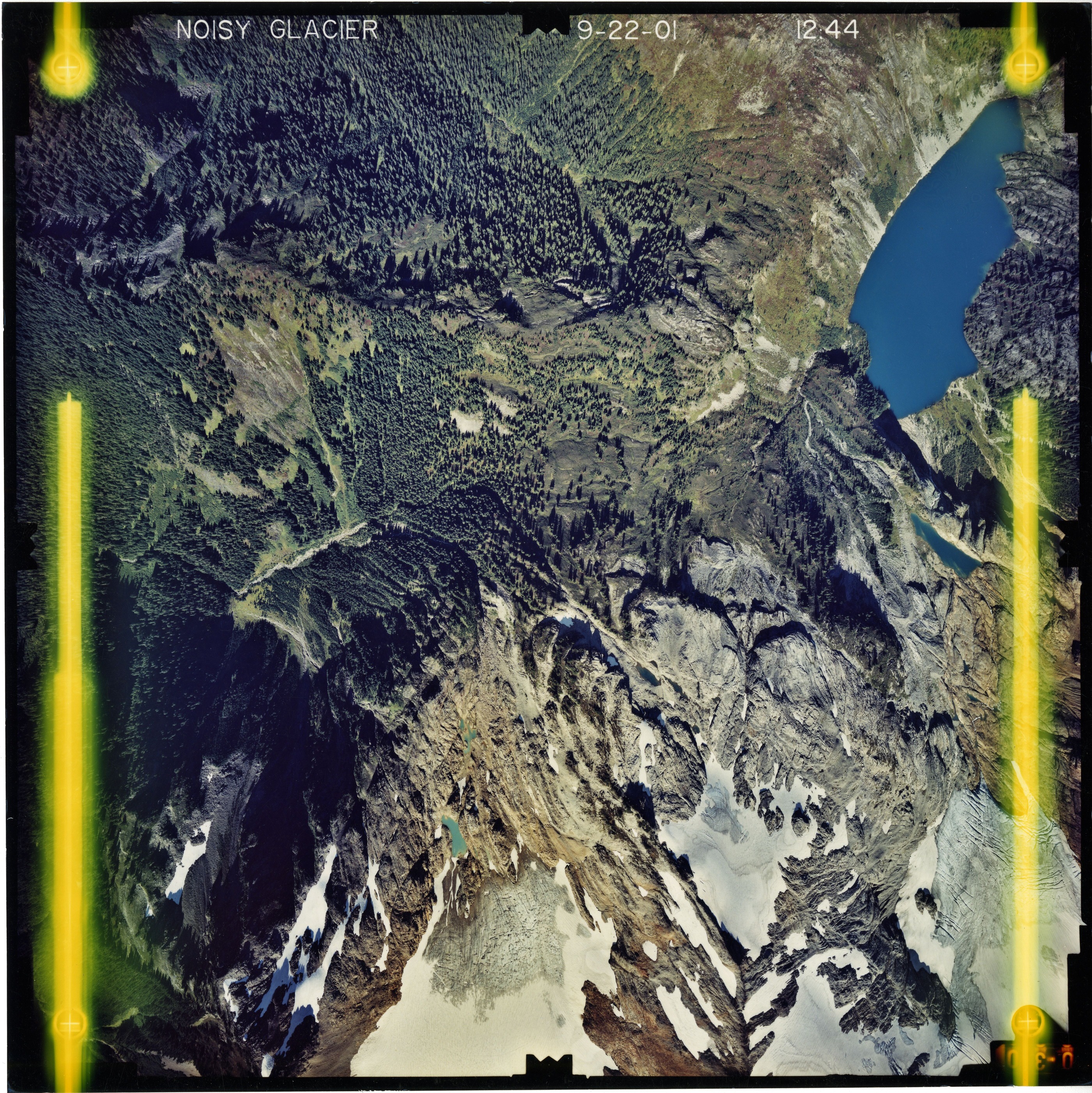 Color aerial photograph of glacier and alpine lake on mountain peak, surrounded by forested valleys.