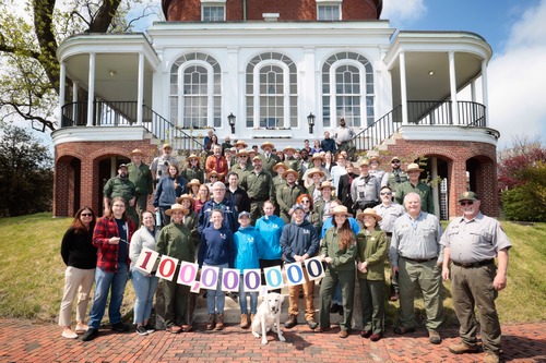 Group of people, some in civilian clothes and others in National Park Service uniforms, standing on the steps of the Commandant's House. A yellow lab sits in the front. People in the front row hold a sign that says "100,000,000"