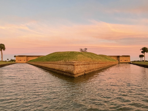A moat surrounds a corner of a low brick wall with a grass mound on top. 