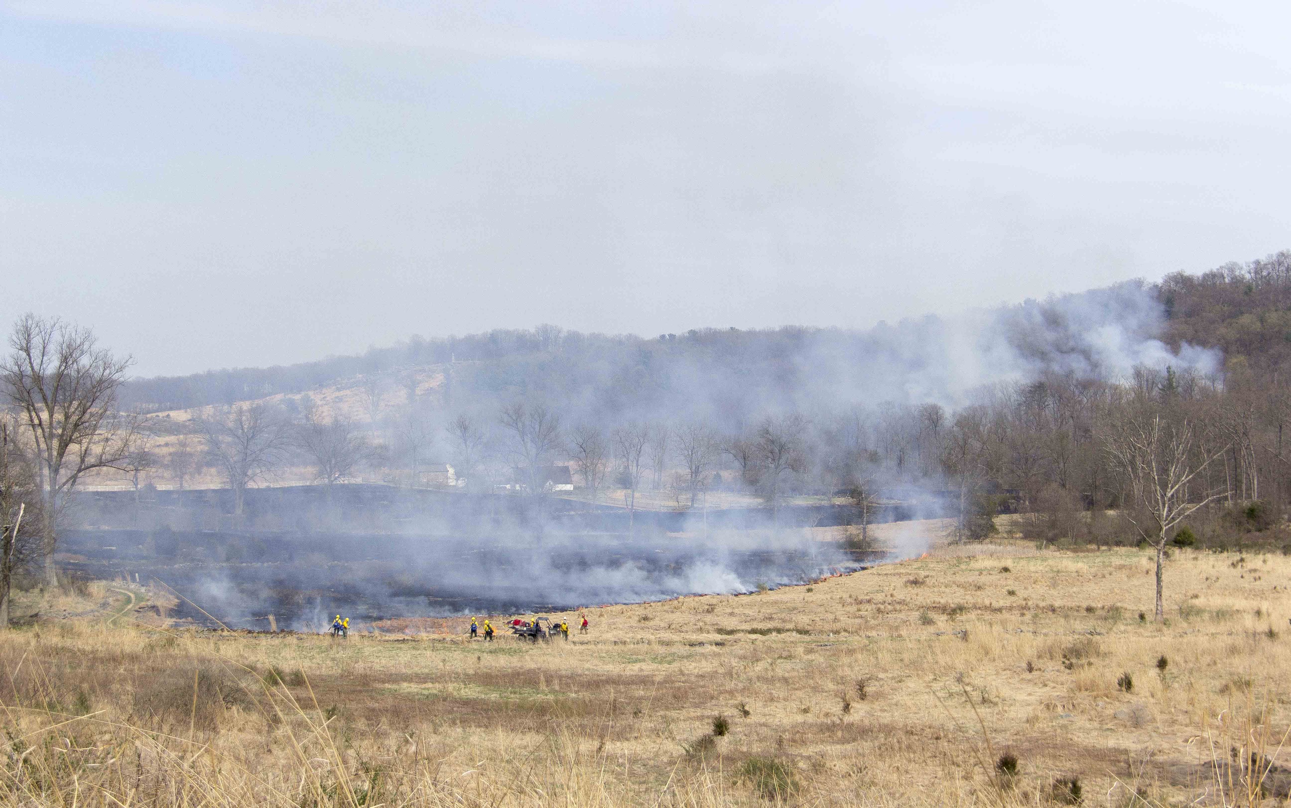 The prescribed fire continues to move south through open fields. Firefighters are in the distance monitoring and caring for the prescribed fire areas. Smoke is coming from the burnt field.