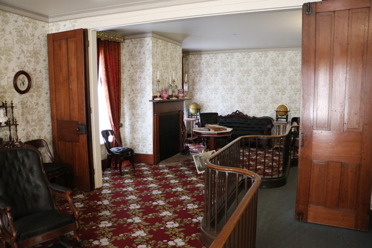 A room with two large wooden folding doors in the middle, red floral carpet, white floral wallpaper, and dark brown and black furniture.