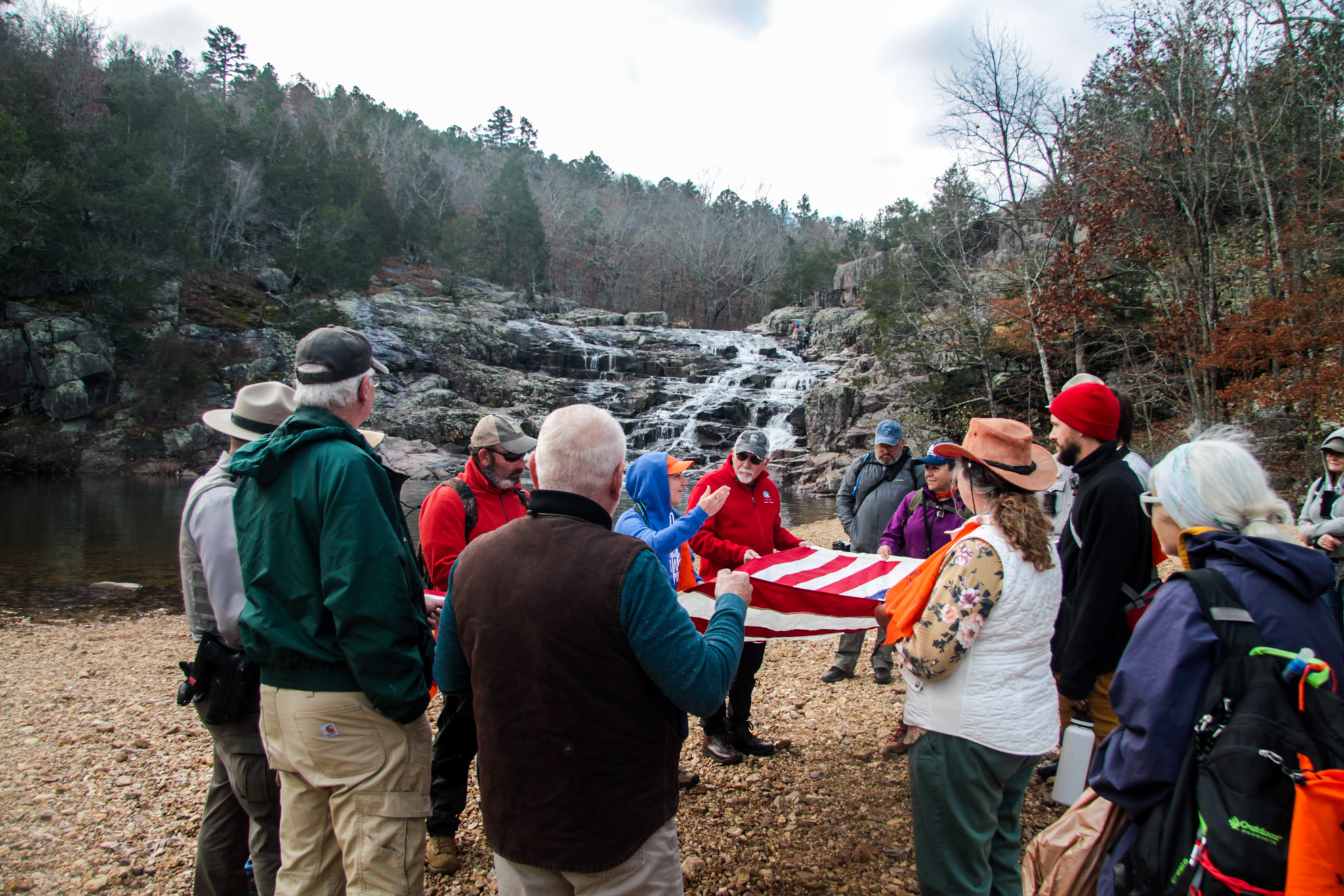 A close-up picture of a group of people gathered around holding the American Flag. In the distance is a cascading waterfall with water flowing. 