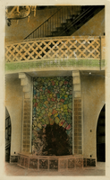 This is an historic black and white photograph hand-tinted from the Scotty's Castle Historic Photograph Collection, Death Valley National Park showcasing interior of two-story room. Staircase with metal handrail running diagonal from ceiling area to second floor perimeter walkway. Across middle is open blockwork walkway railing. Tile work at base of openwork is tinted green. Below is elaborate rock faced fountain framed in tile with tiled pond at bottom. Rocks individually tinted various colors. Tiles along edge tinted yellow. Basin tiles tinted pink. Arched openings either side fountain. Tiled floor.
