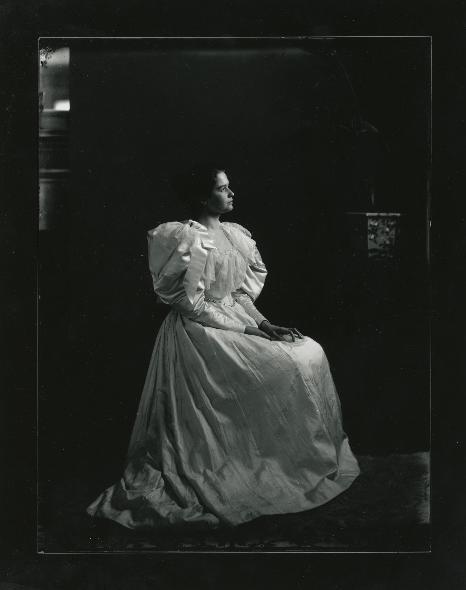 A young white woman dressed in a puffed-sleeve dress sits in angled to her left.