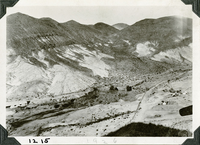 This is an historic black and white photograph from the Scotty's Castle Historic Photograph Collection, Death Valley National Park of barn on the left edge. A dense area of vegetation marks a spring. A dirt road runs along the base of barren, steep desert mountains. Fence line and road run up the length of the wash on right side. Scattering of tents and small structures to right of road. Plane prop on right edge.
