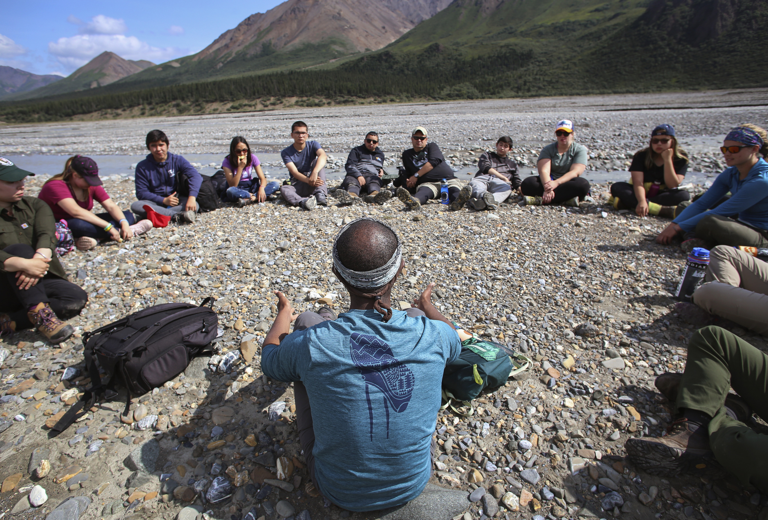 teenagers sitting in a circle on the ground near a river, listening to a man speak