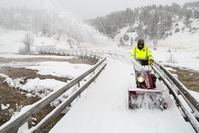 A uniformed man in winter clothing pushes a snowblower down a boardwalk clearing the snow.