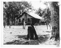 A woman in a dress standing beside a fence outside a wooden house built in a field.