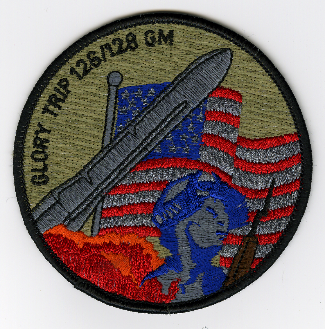 Olive green with black border. American flag in center, red, grey and blue colors. Minuteman III missile diagonally from bottom left corner going over blue field of flag to top right corner. Orange cloud at bottom, representing missile launch. Figure in bottom right with musket representing minute man of the 18th century.
