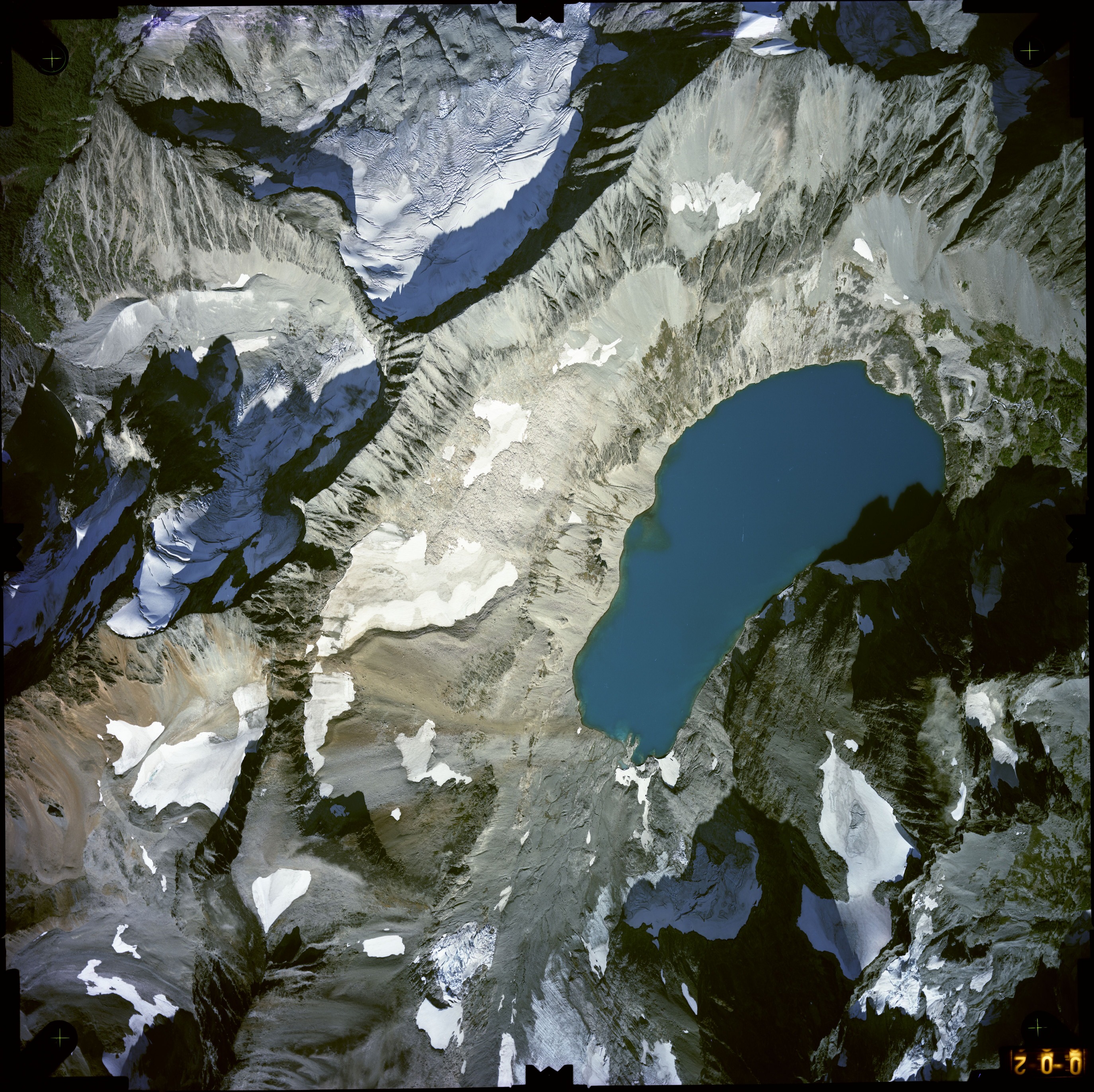 Color aerial photograph of glacier, snowfields, and alpine lake on mountain peak with forested valleys below.
