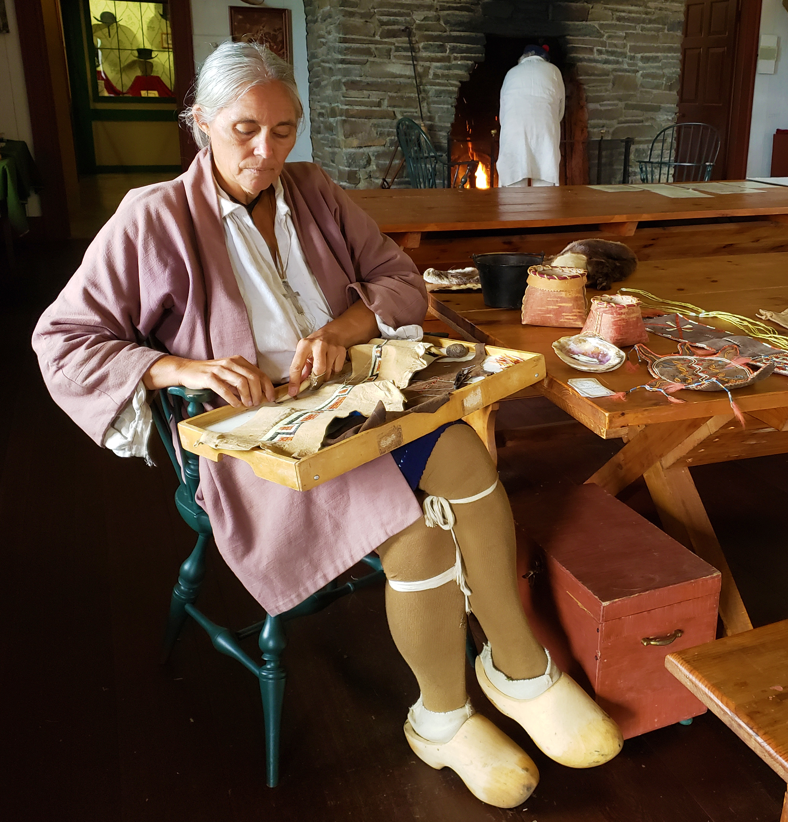 A person in historic clothing seated next to a table with a tray holding a piece of leather with porcupine quill work in progress.