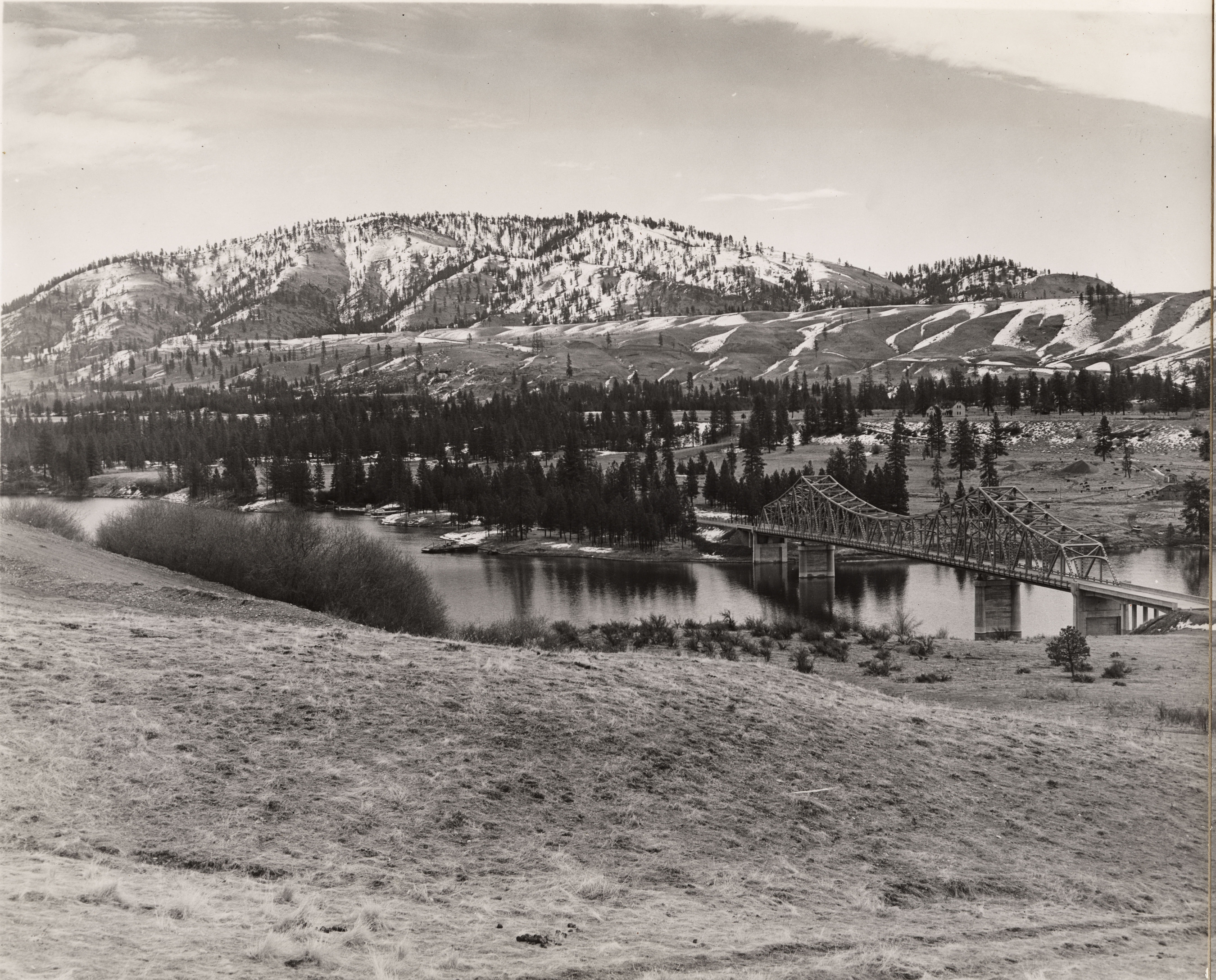Black and white photograph of metal bridge across a river with snowy mountains rising above