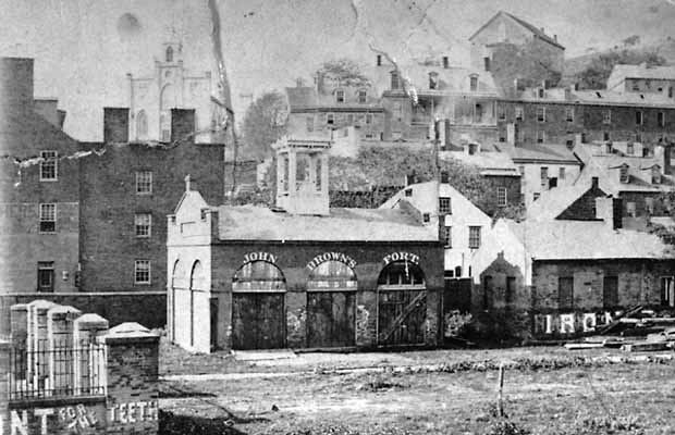 1882-1886 photo of John Brown Fort. The building to the right of the fort was the former Armory superintendent's office. Advertisements painted on buildings and walls were aimed at rail travelers passing through Harpers Ferry. Year: 1885. Image Credit: Historic Photo Collection, Harpers Ferry NHP.