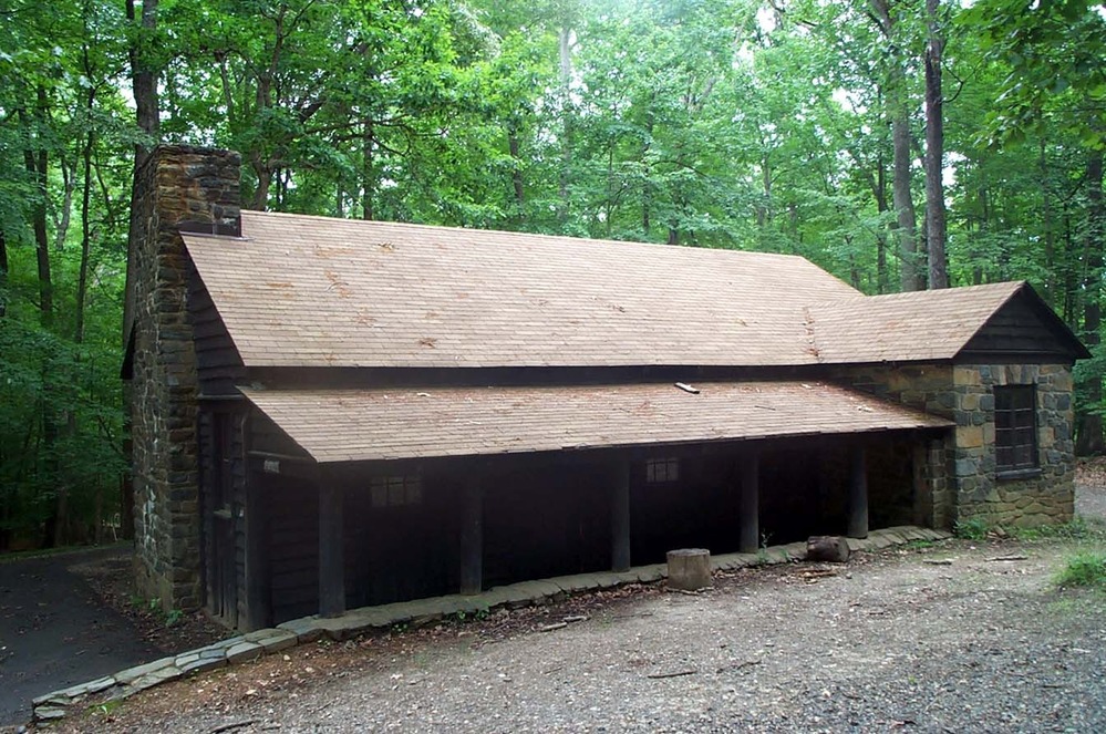 Wooden building with a stone fireplace and porch with a paved walkway beside it at the edge of the green forest