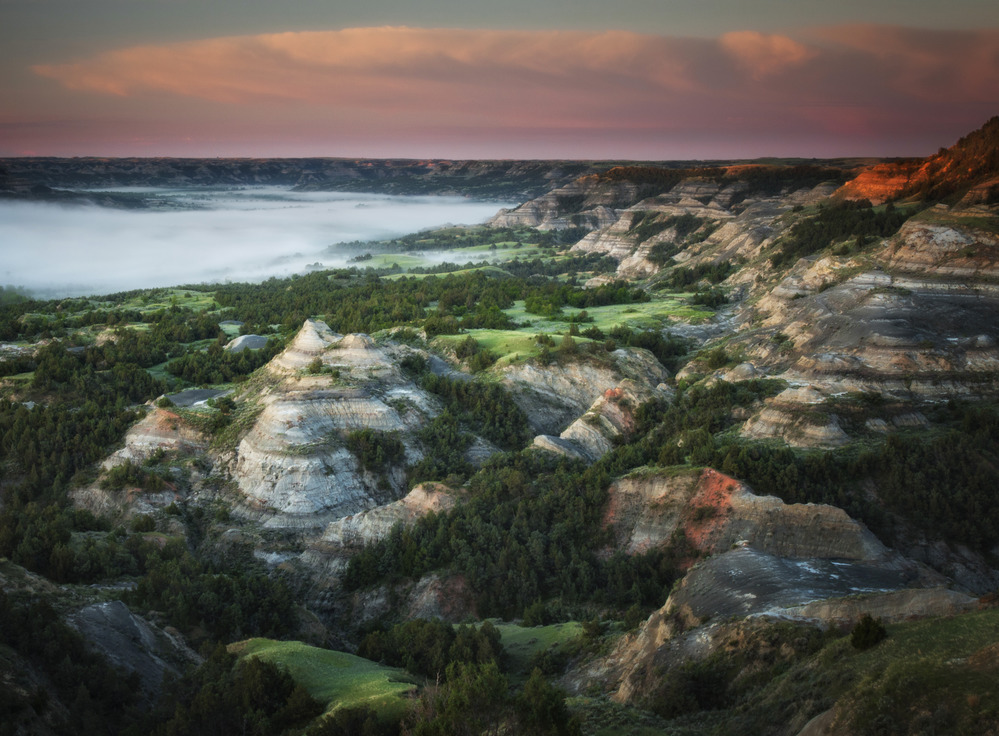 Vibrant pink sky over barren buttes and valley of lush green forest. 